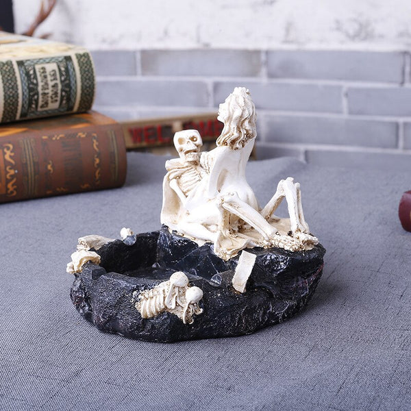 Cendrier tête de mort en résine noire avec femme et squelette ivoire sculptés, cavité entourée d’ossements